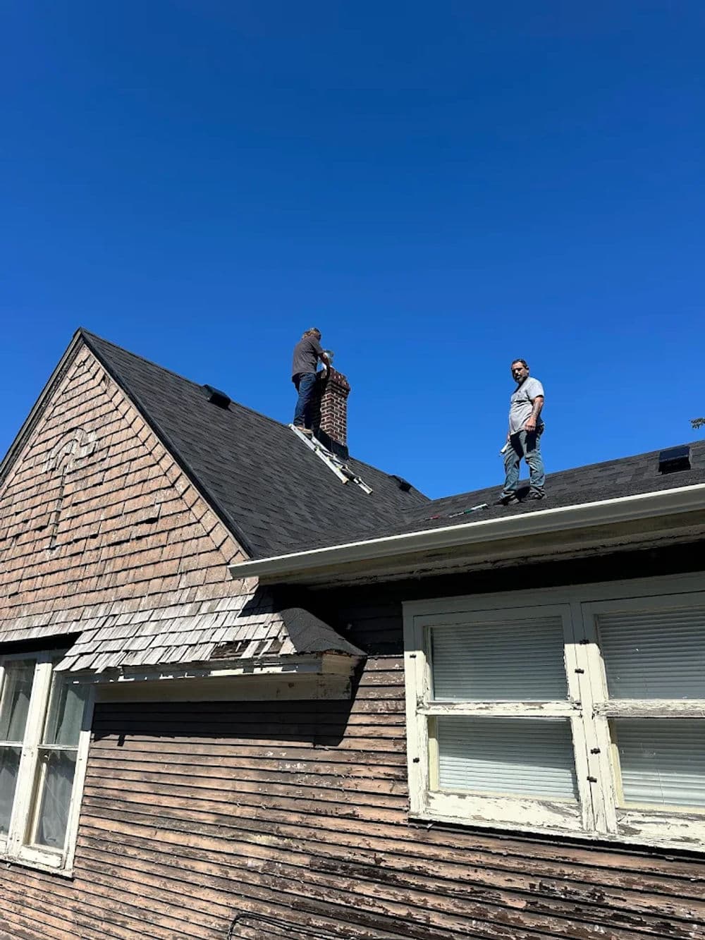 Two workers repairing a chimney on a worn wooden roof under a clear blue sky.